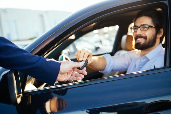 A person hands car keys to someone sitting inside a car.