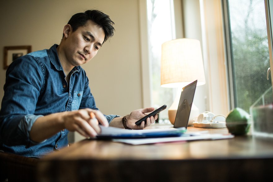 Adult sitting at desk reviews paperwork in front of laptop and with smartphone in hand.