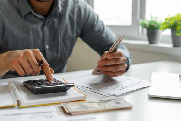 Person using calculator while holding cash and looking at papers on a desk.
