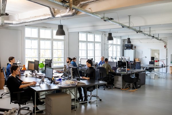 Workers sitting at desks in trendy office setting with large windows and high ceilings.