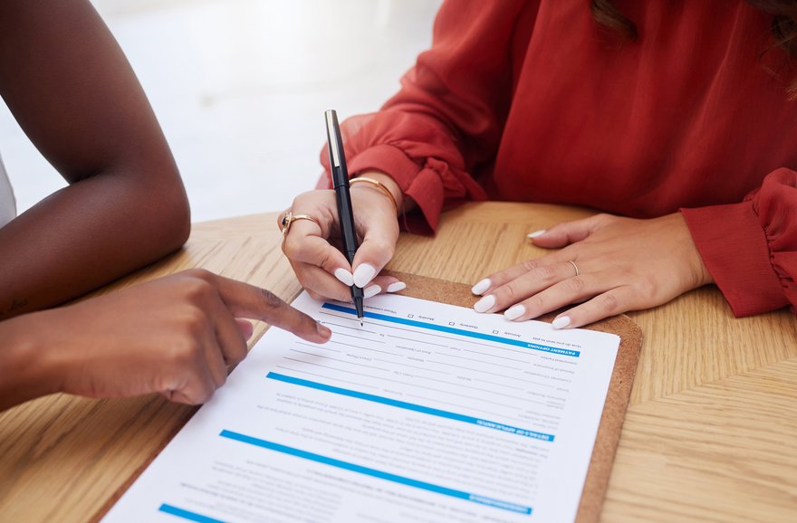 Close-up shot of a document on a table, with one person pointing to where the other person should sign it.